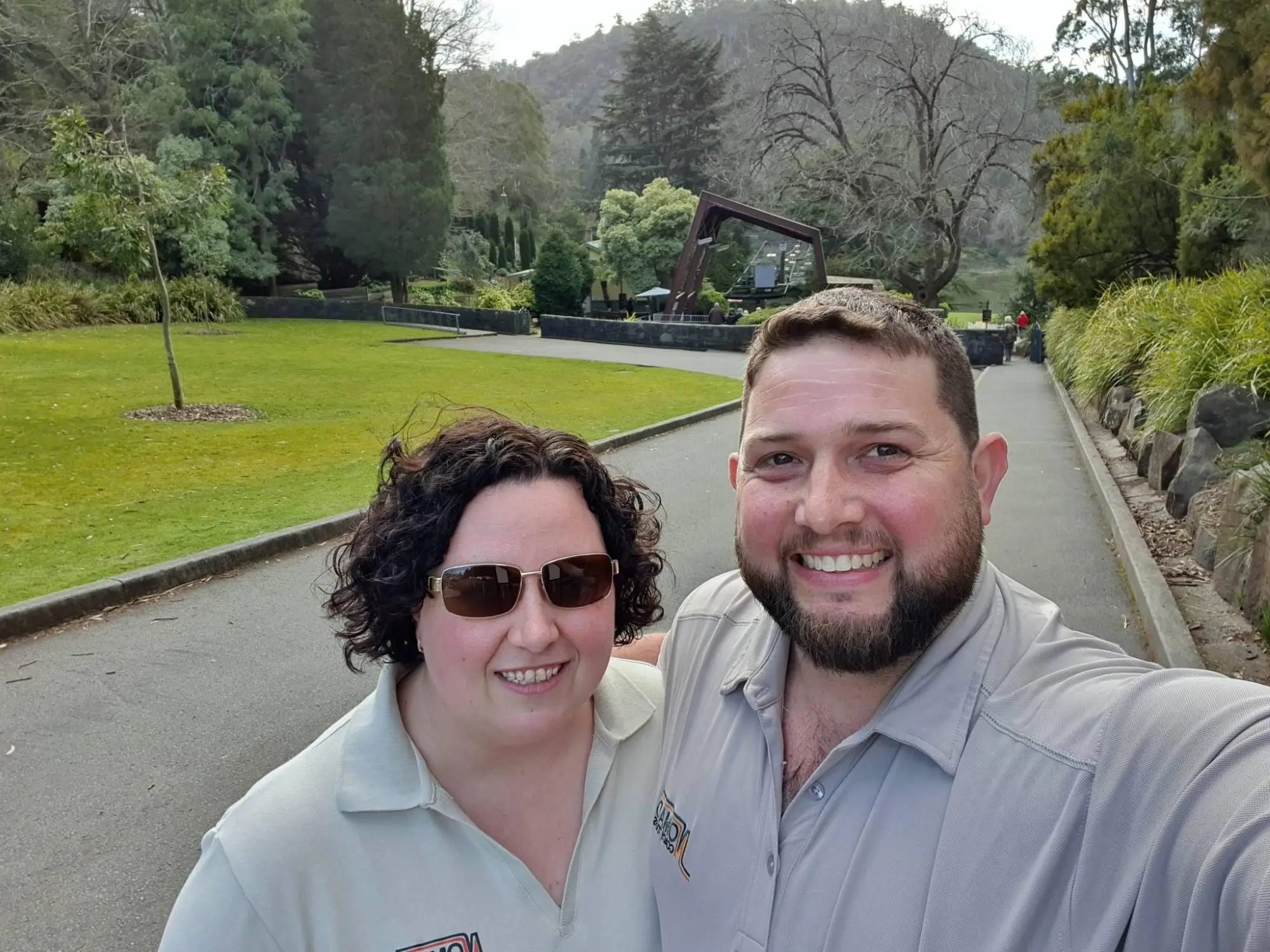 A man and a woman smile for a selfie on a paved path surrounded by greenery. Both are wearing gray shirts. Trees and a small wooden structure are in the background. It's a sunny day.