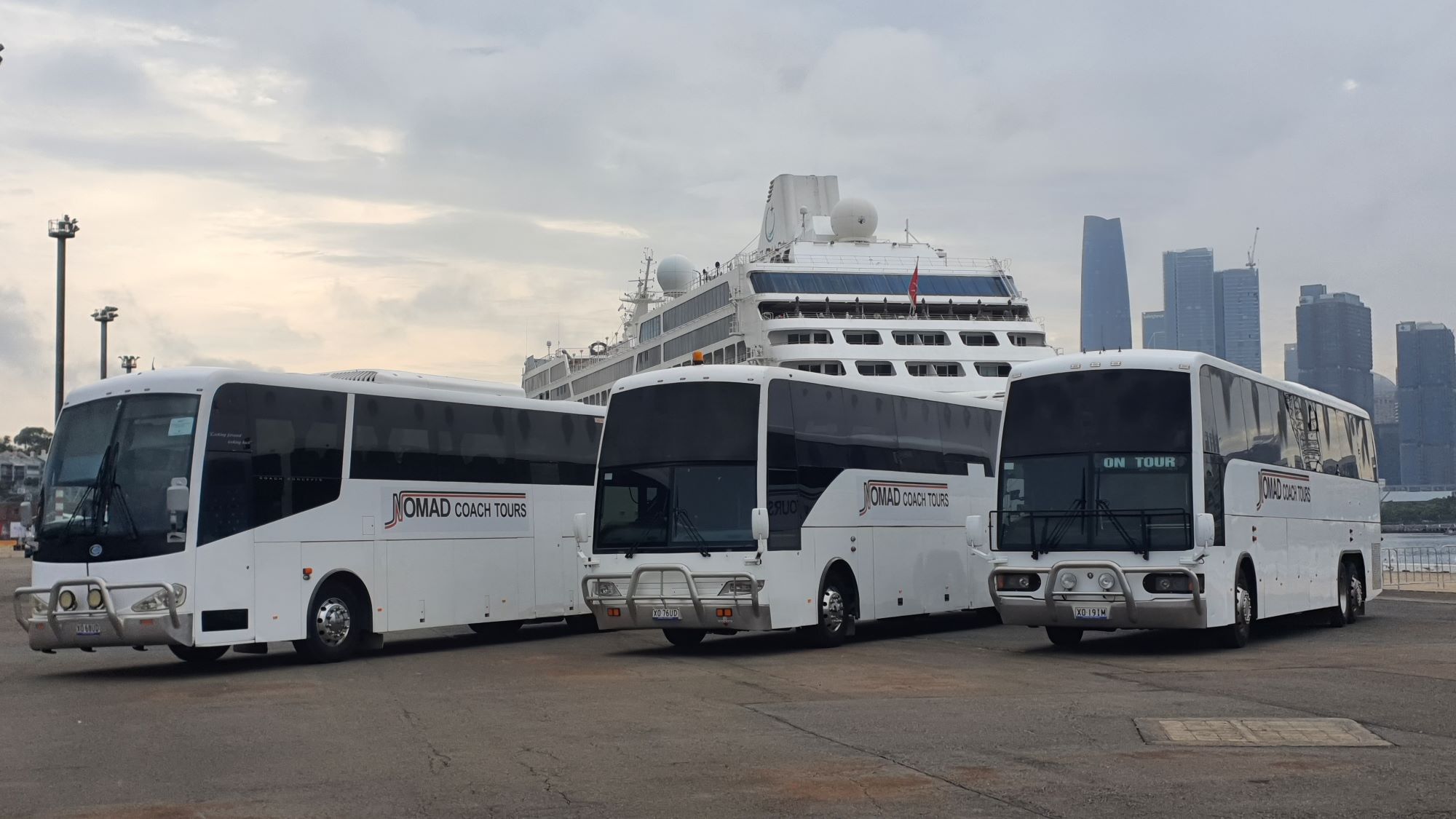 Three white tour buses parked in front of a large cruise ship. The buses have "Coach Tours" logos, and a city skyline with tall buildings is visible in the background under a cloudy sky.