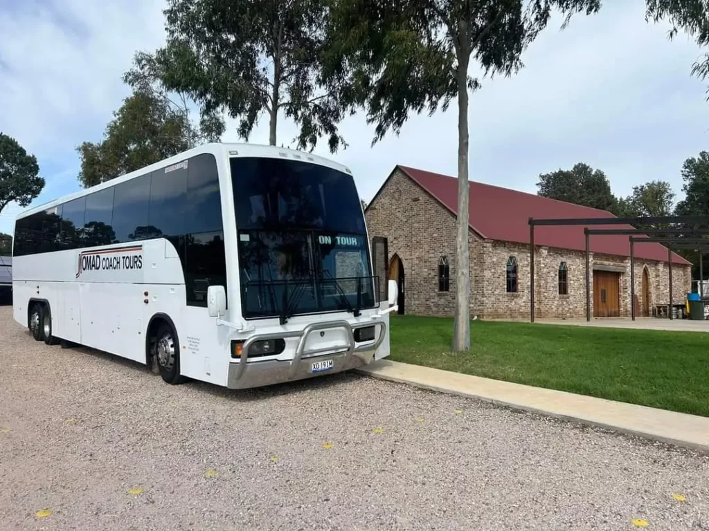 A white tour bus labeled "On Tour" represents Nomad Coach Tours services as it rests on a gravel path beside a stone building with a red roof. The scene is embraced by trees and grass, creating a peaceful, scenic atmosphere.