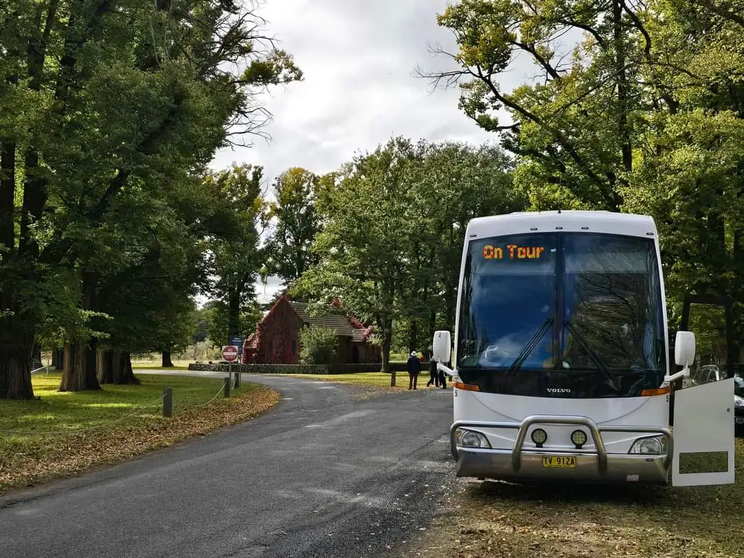 A white tour bus with the sign "On Tour" is parked on a road lined with green trees. A brick building with a red roof is visible in the background. The sky is cloudy, and the road is bordered by grass and leaves.