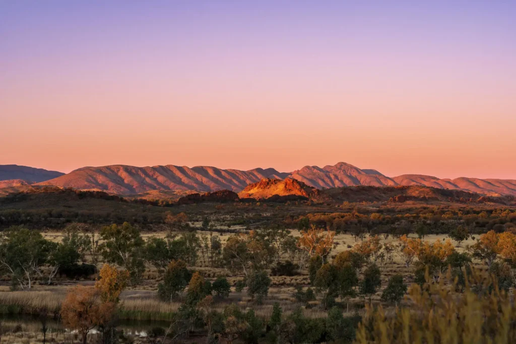 Arid landscape at sunset with a range of mountains in the background, glowing orange and pink. The foreground features sparse trees and dry grass under a clear sky that fades from purple to blue.