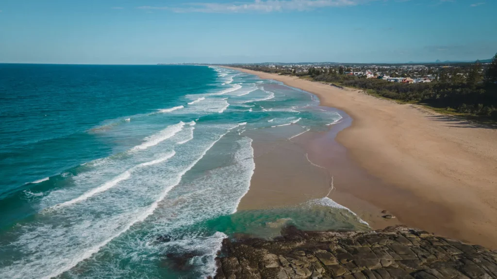 A wide sandy beach with gentle waves rolling in from a turquoise ocean, bordered by a rocky shoreline in the foreground and a distant coastal town under a clear blue sky.