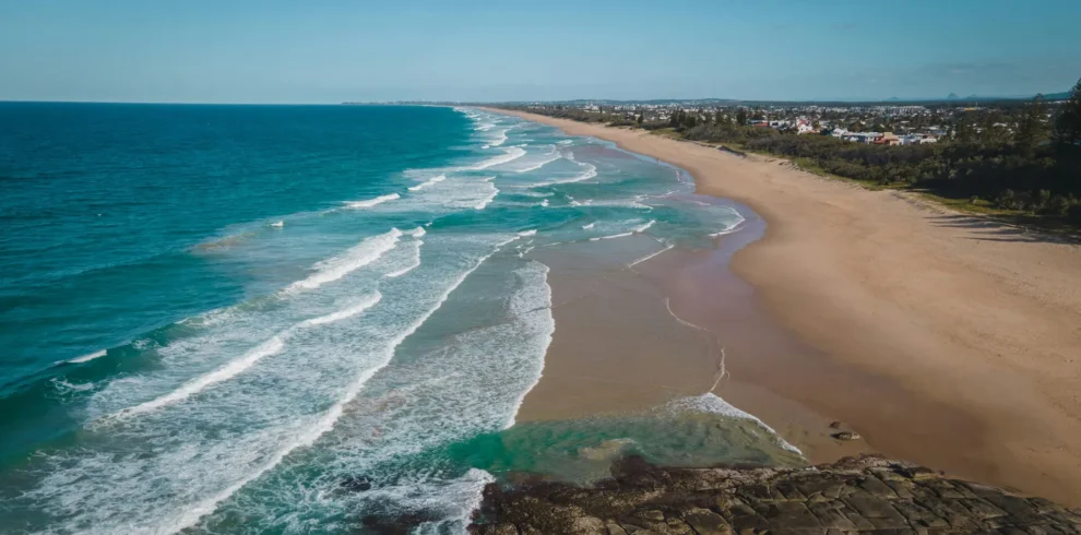 A wide sandy beach with gentle waves rolling in from a turquoise ocean, bordered by a rocky shoreline in the foreground and a distant coastal town under a clear blue sky.