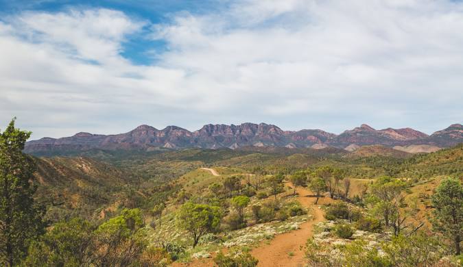A scenic landscape view of a dirt trail winding through green shrubland and rocky hills, with a mountain range and blue sky with scattered clouds in the background.