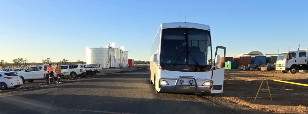 A white bus with an open door is parked on a paved road at a worksite, with industrial tanks, vehicles, and workers in safety vests visible in the background under a clear blue sky.