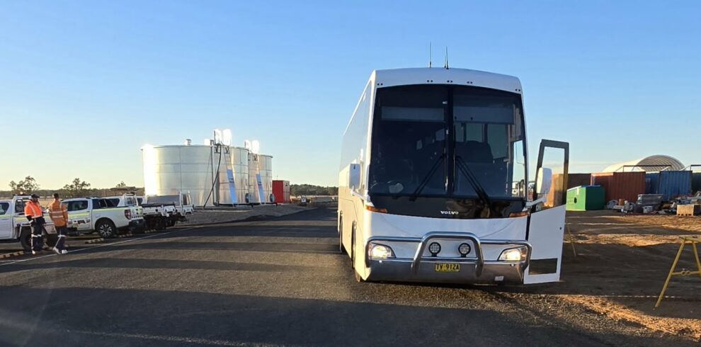 A white bus with an open door is parked on a paved road at a worksite, with industrial tanks, vehicles, and workers in safety vests visible in the background under a clear blue sky.