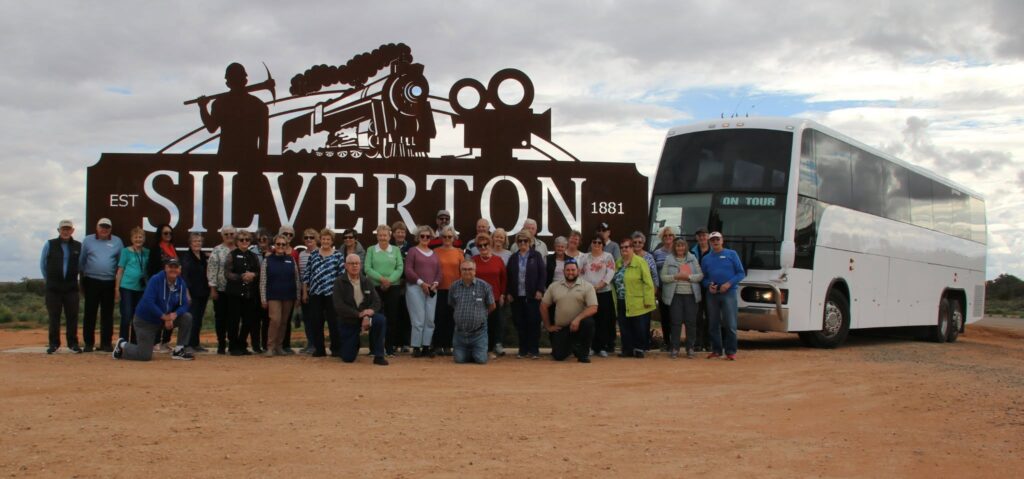 A large group of people poses in front of a "Silverton" sign featuring a train silhouette, with a white tour bus parked beside them on a dirt road under a cloudy sky.