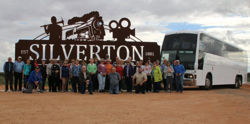 A large group of people poses in front of a "Silverton" sign featuring a train silhouette, with a white tour bus parked beside them on a dirt road under a cloudy sky.