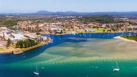 Aerial view of a coastal town with buildings, greenery, a blue river merging with the sea, boats anchored in the water, and distant mountains visible under a clear sky.